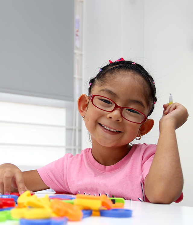 A young girl with glasses and a hairband smiles at the camera while sitting at a table with colorful toys in front of her.